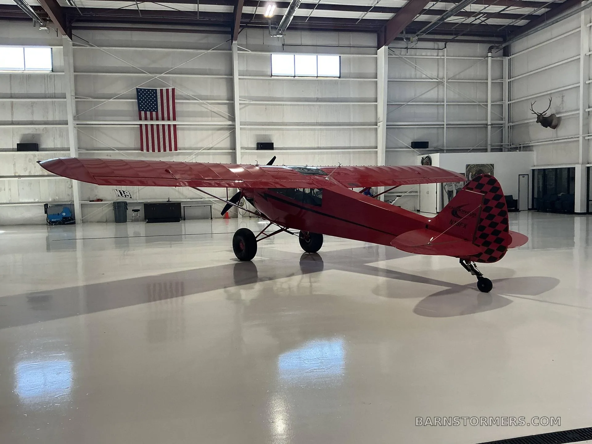 A red Bushmaster aircraft with a checkered tail is parked in a hangar, viewed from the rear-right.