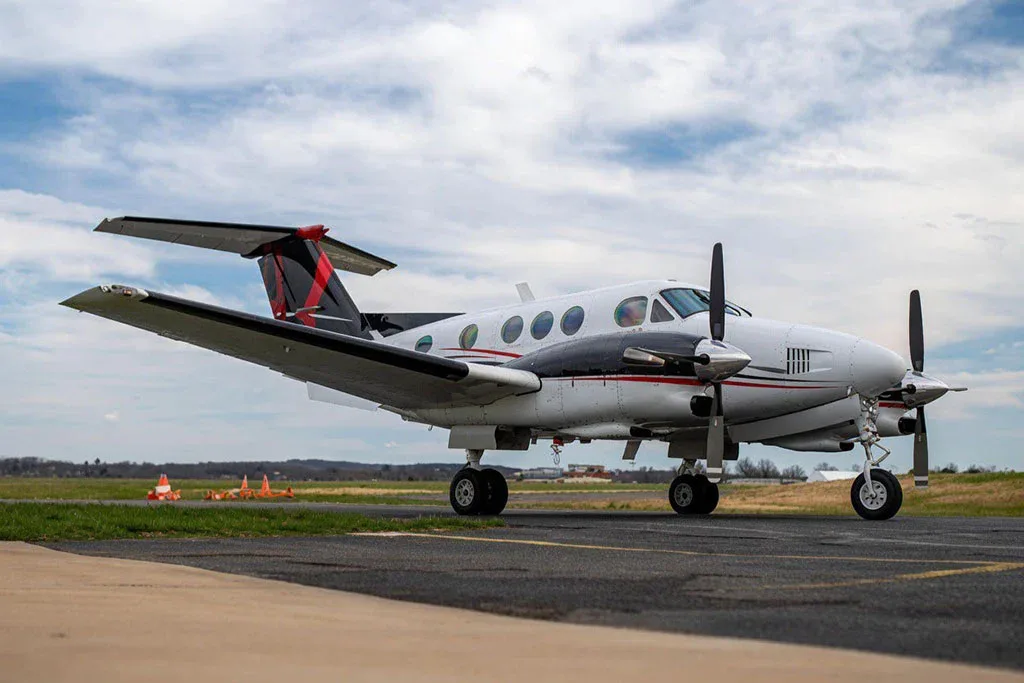 A white and black Beechcraft King Air with red accents on the tail, parked on an airport tarmac under a cloudy sky.