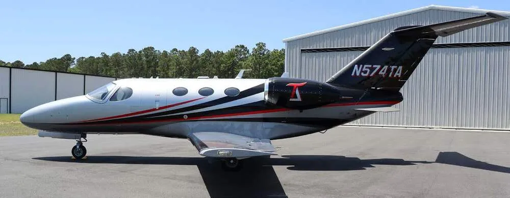 A silver, black, and red Cessna Citation Mustang jet, tail number N574TA, parked on an tarmac next to a hangar.