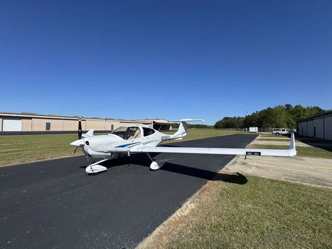 A white Diamond DA40 aircraft with blue stripes is parked on an airport ramp under a clear blue sky.