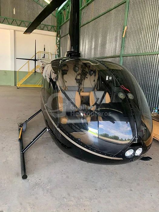 A close-up front view of a black Robinson R66 Turbine helicopter showing its tan interior seats through the clear canopy, inside a hangar.