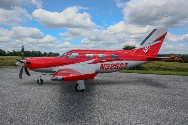 A red and white Piper M600 SLS aircraft with registration N325ST is parked on an asphalt ramp under a partly cloudy sky, viewed from the left side.