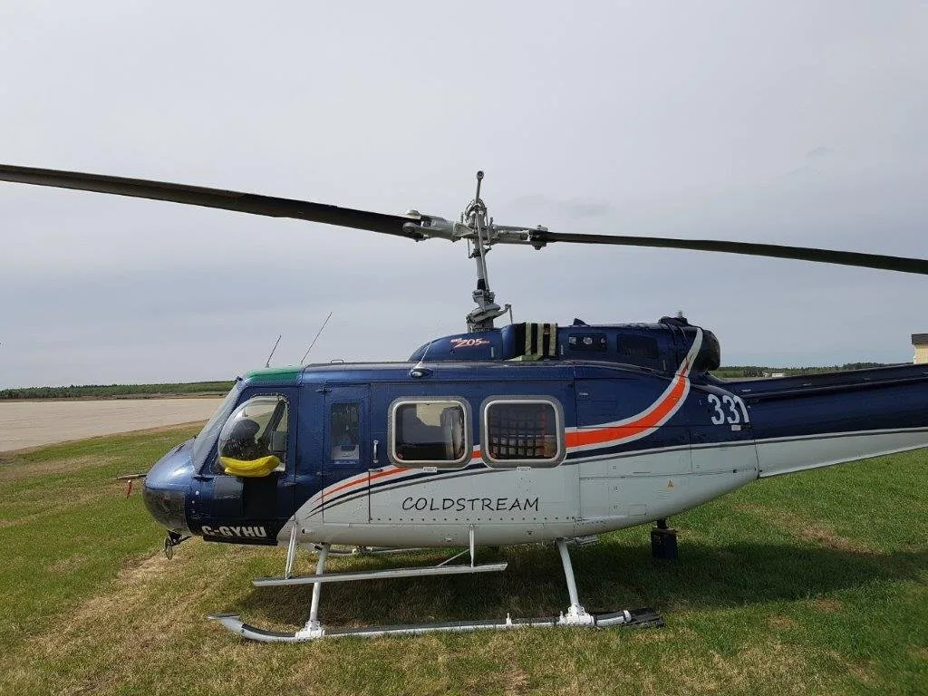 A blue and white Bell 205 helicopter with orange stripes, registration C-GYHU, parked on grass next to an airfield.