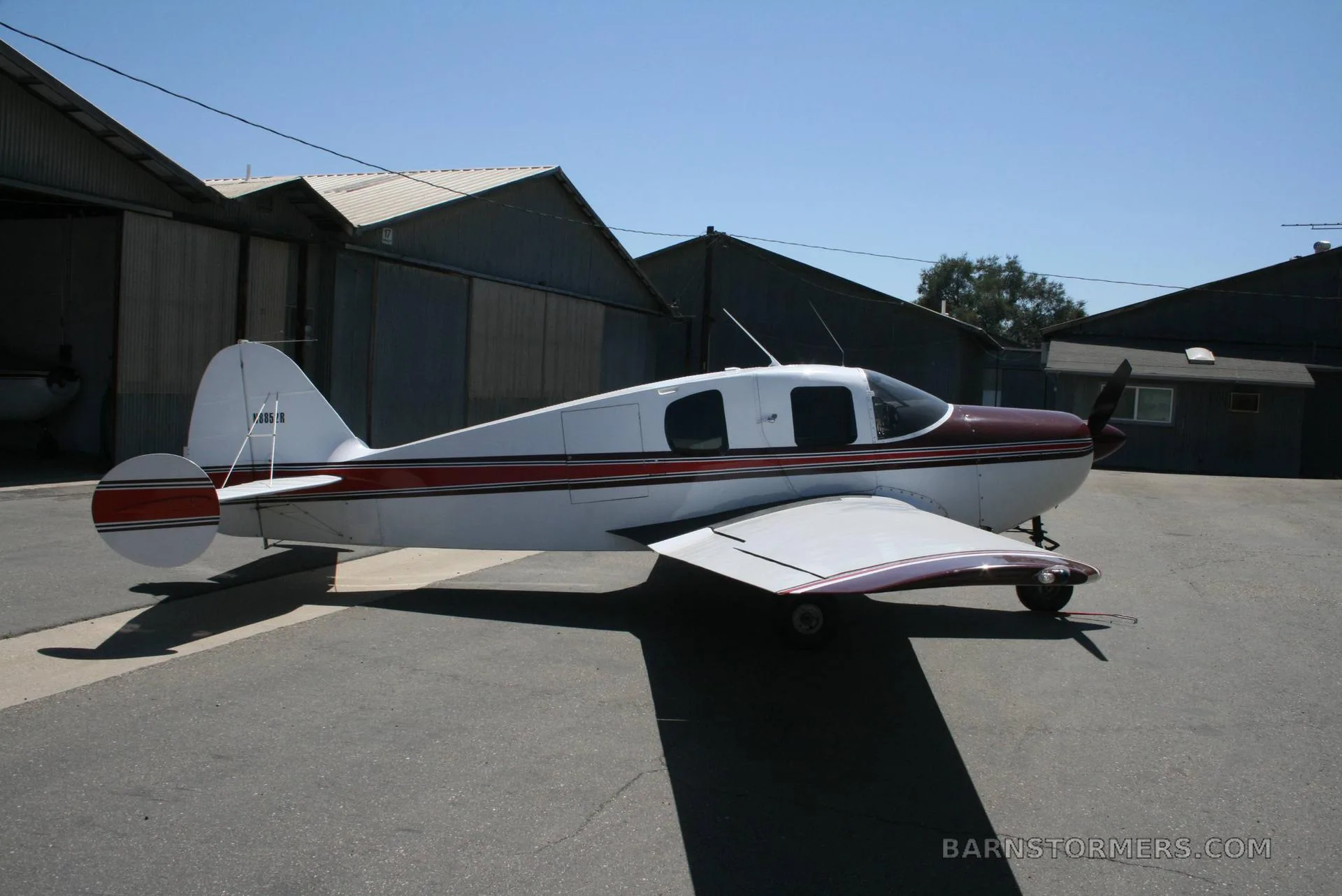 A white and maroon single-engine aircraft, tail number N8352R, parked on an asphalt apron with hangars in the background under a clear blue sky.