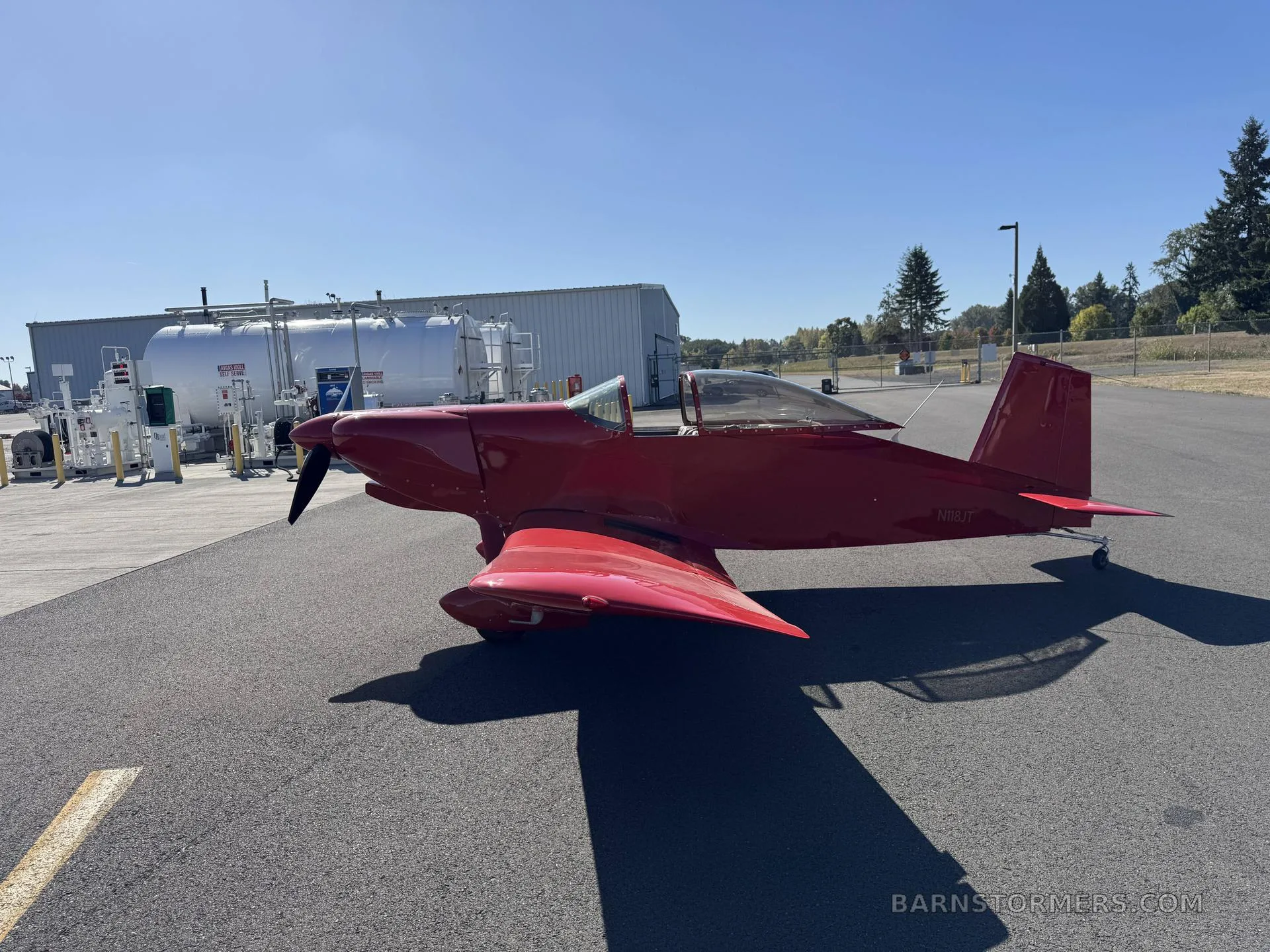 A bright red single-engine experimental aircraft (N18JT) is parked on an airfield with fuel tanks and a building in the background under a clear blue sky.