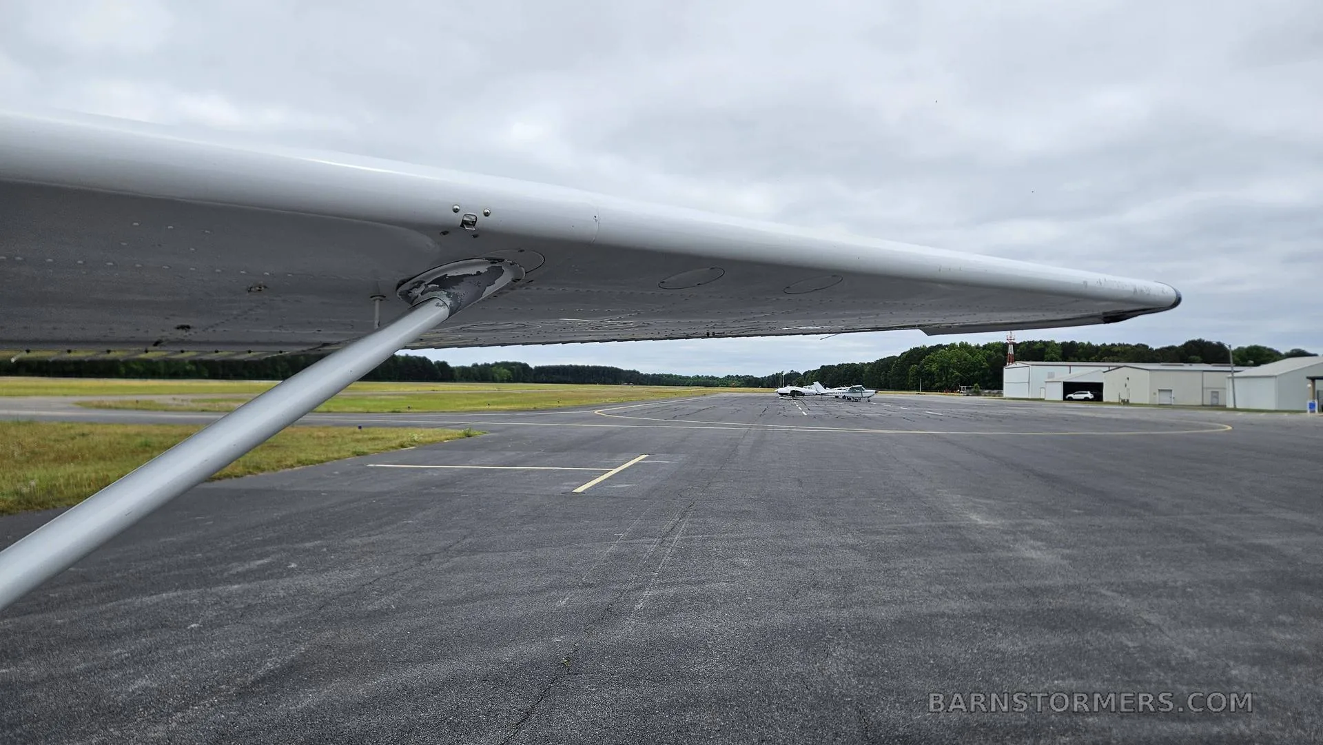 A view from under the wing of an aircraft, looking down a tarmac towards a hangar and another small plane in the distance.