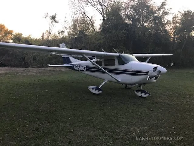 A white and blue Cessna 182 aircraft is parked on a grassy field at sunset.