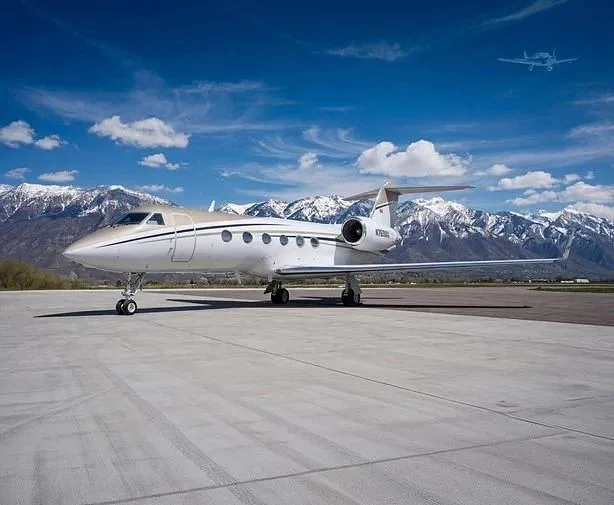 A white and gold Gulfstream G550 business jet, N769MM, is parked on an airfield with snow-capped mountains under a blue sky.