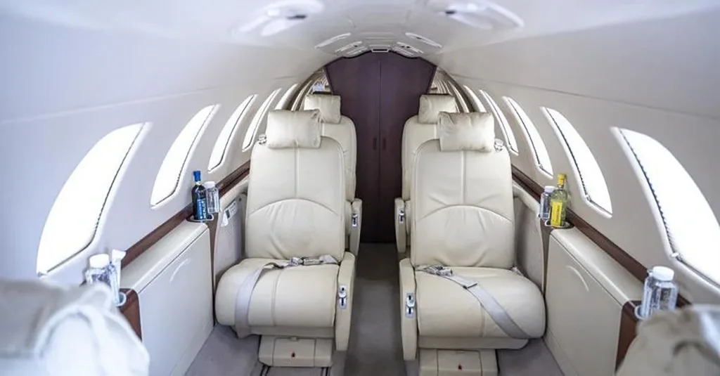 A view down the aisle of a private jet cabin, showing light beige leather seats with bottles on side tables.