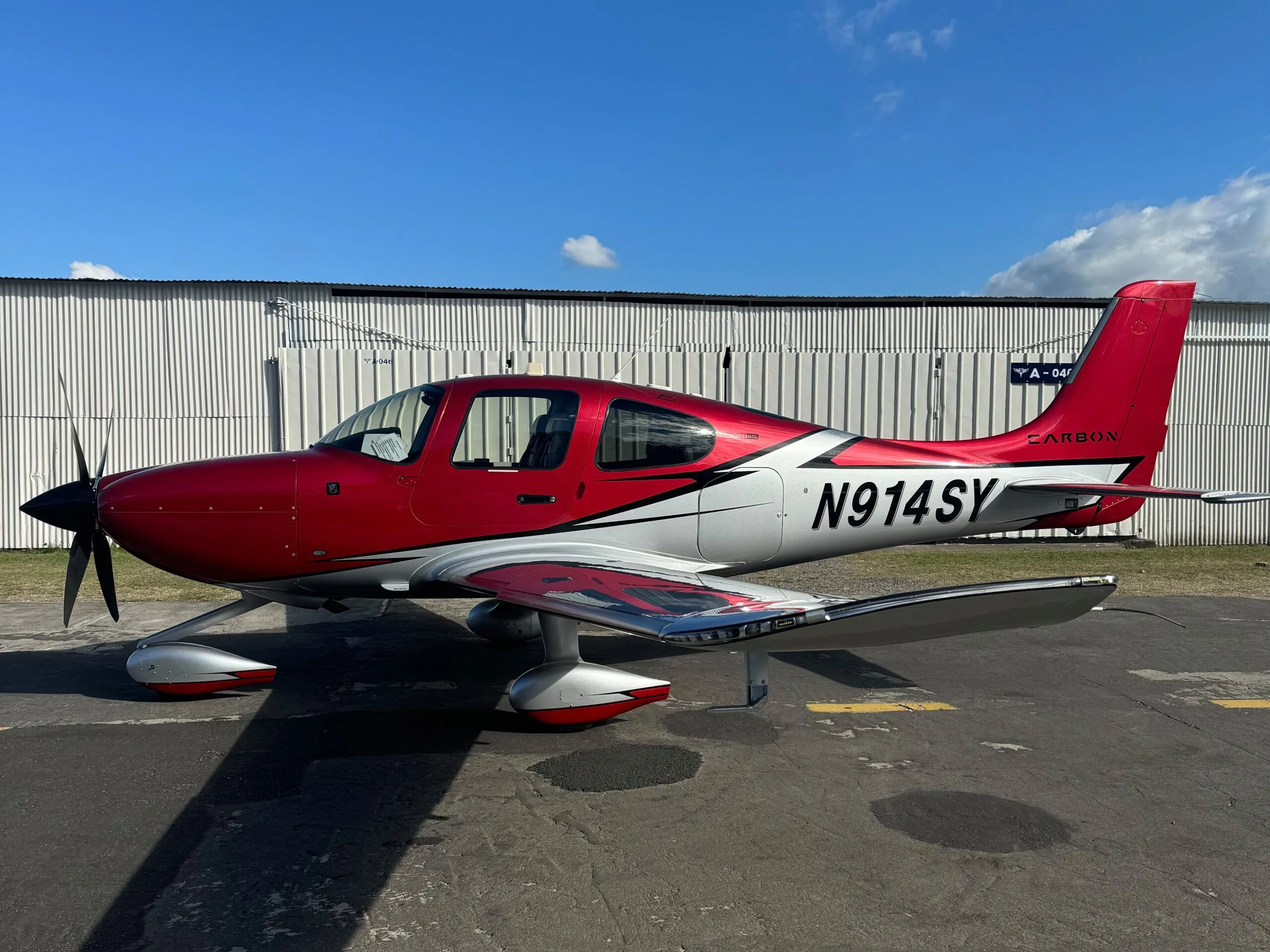 A red and silver Cirrus SR22 aircraft with registration N914SY parked on an asphalt apron in front of a white hangar.
