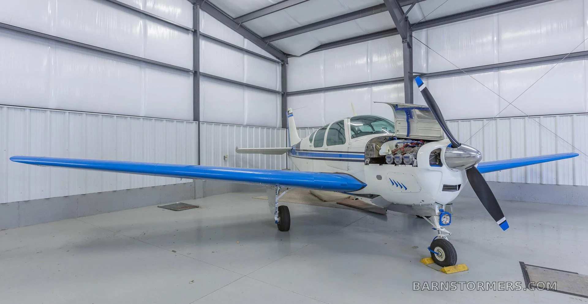 A white and blue single-engine aircraft with its engine cowl open, parked inside a hangar.