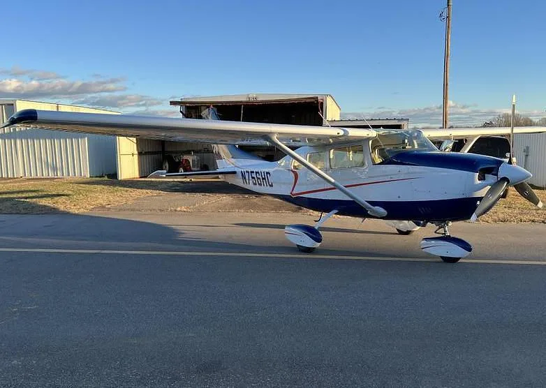 A white and blue Cessna 182, N756HC, parked on an asphalt ramp with hangars in the background under a clear sky.