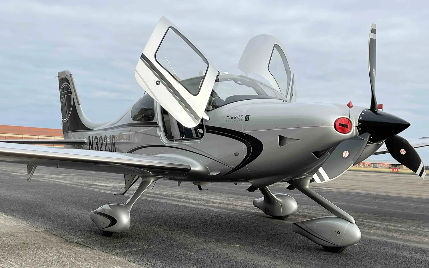 A silver Cirrus SR22 aircraft with registration N322JR and open doors is on an airfield under a cloudy sky.
