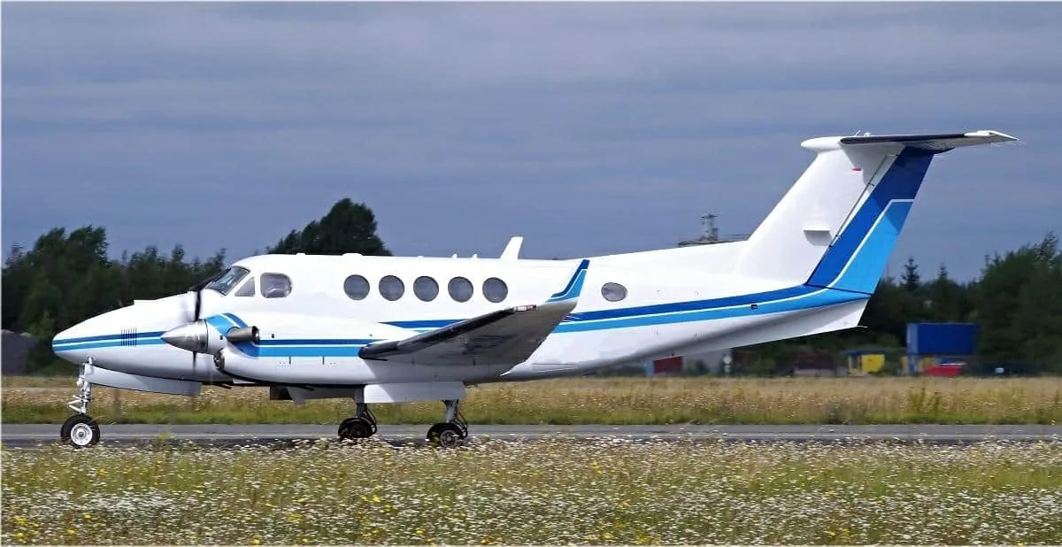 A white and blue Beechcraft King Air turboprop aircraft is taxiing on a runway next to a field of wildflowers.