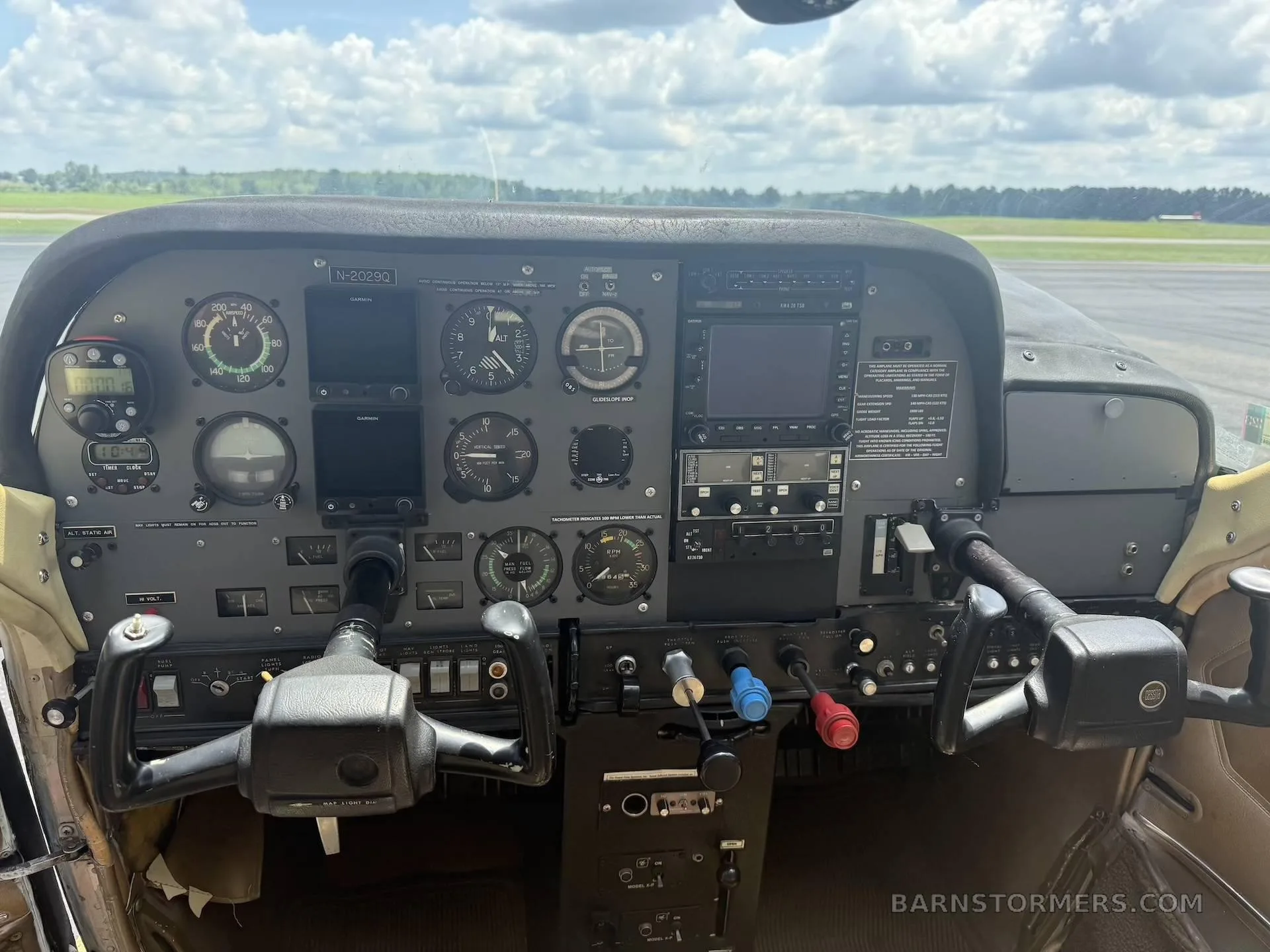 The instrument panel and yokes of a small aircraft cockpit, with a view of the runway and trees outside.