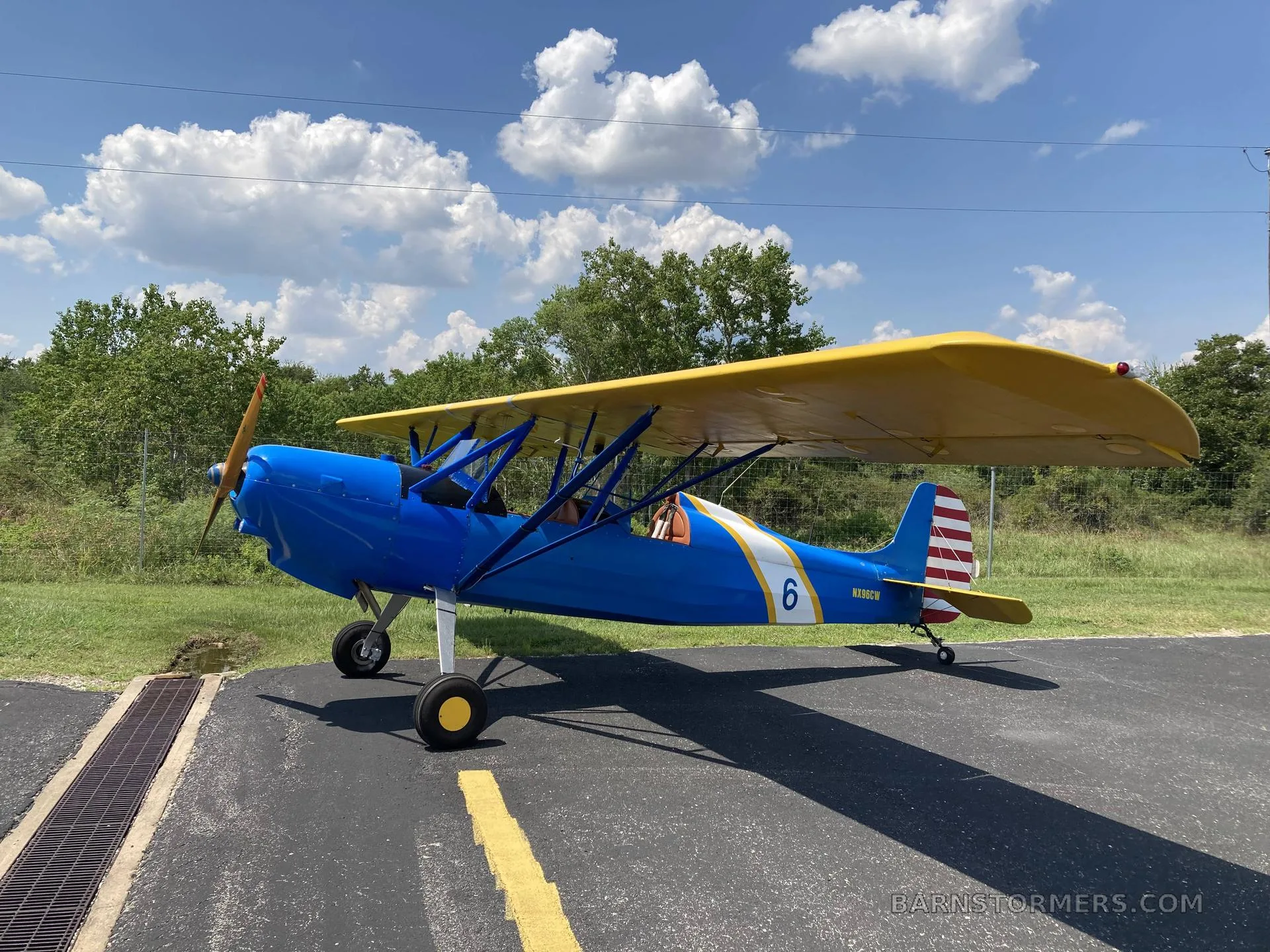 A blue and yellow biplane with '6' on the side, parked on an asphalt runway under a blue sky with clouds.