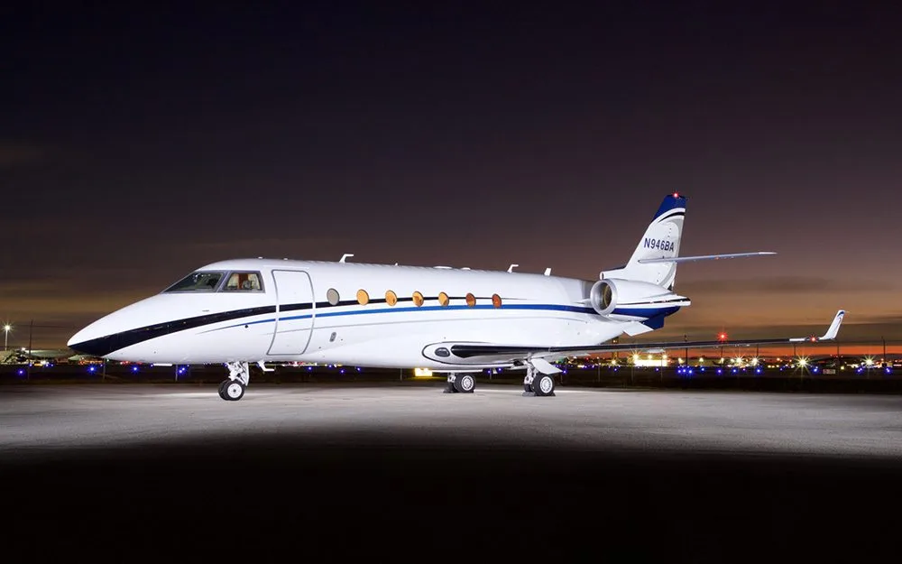 A white business jet with blue and black stripes, registration N946BA, parked on an airfield at night.