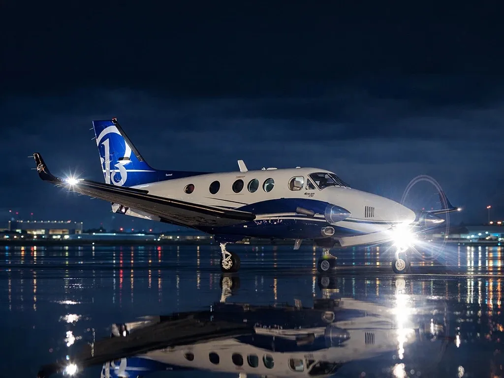 A white and blue Beechcraft King Air aircraft with a "15" logo on its tail sits on a wet tarmac at night, reflecting city lights.