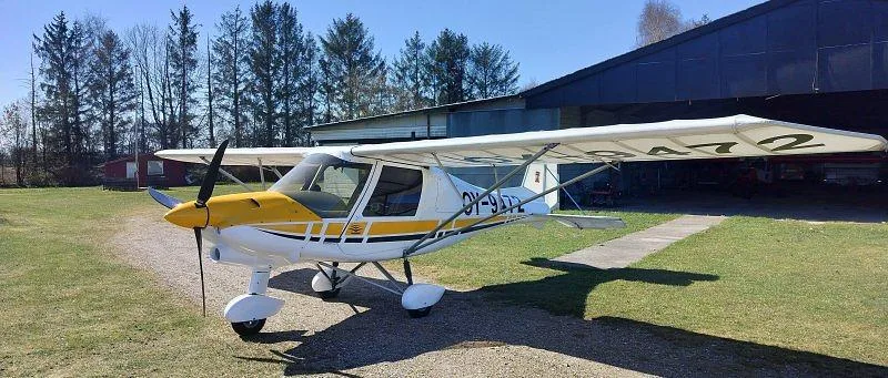 A white and yellow light sport aircraft, registration OY-9472, parked on grass in front of a hangar on a sunny day.