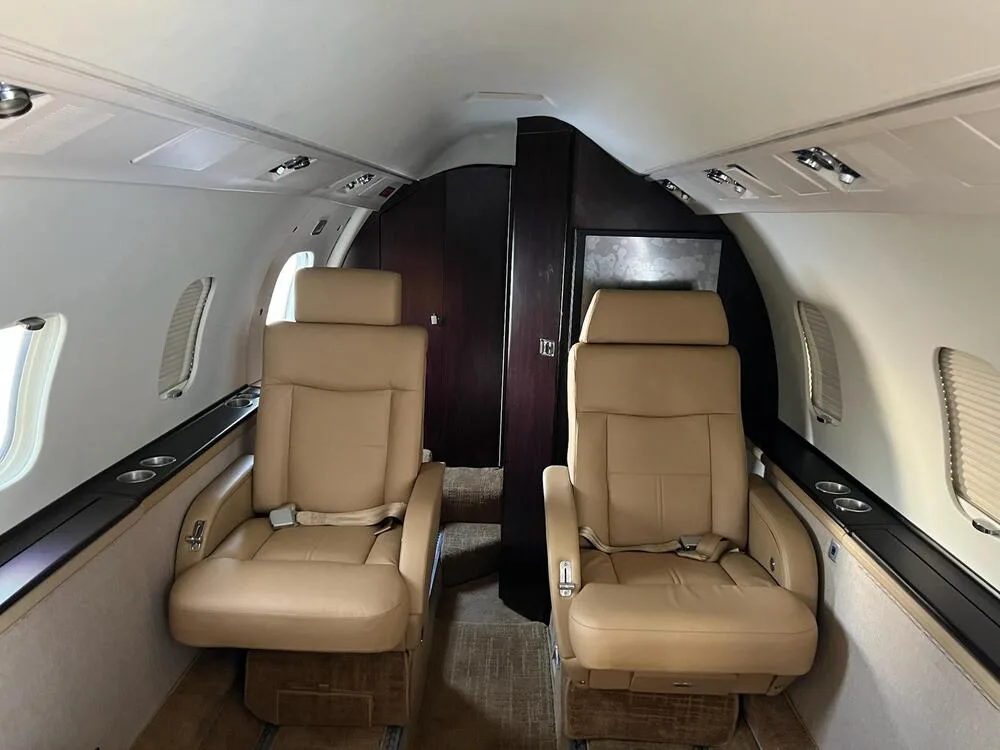 View of two tan leather passenger seats in the aircraft cabin with dark wood paneling and overhead lighting.