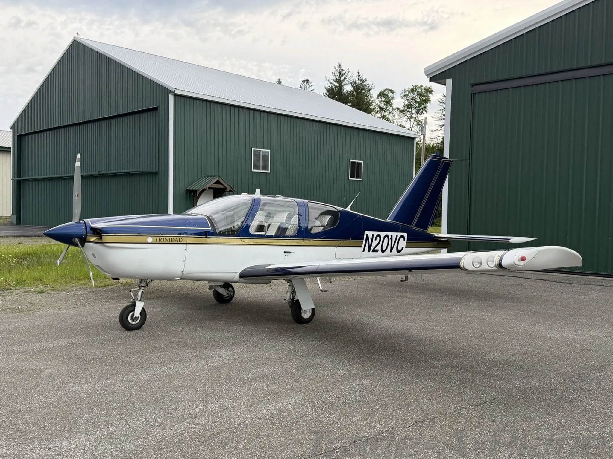 A side view of the N20VC Socata Trinidad TB20 aircraft, painted white and blue with gold accents, parked on an airfield next to green hangars.