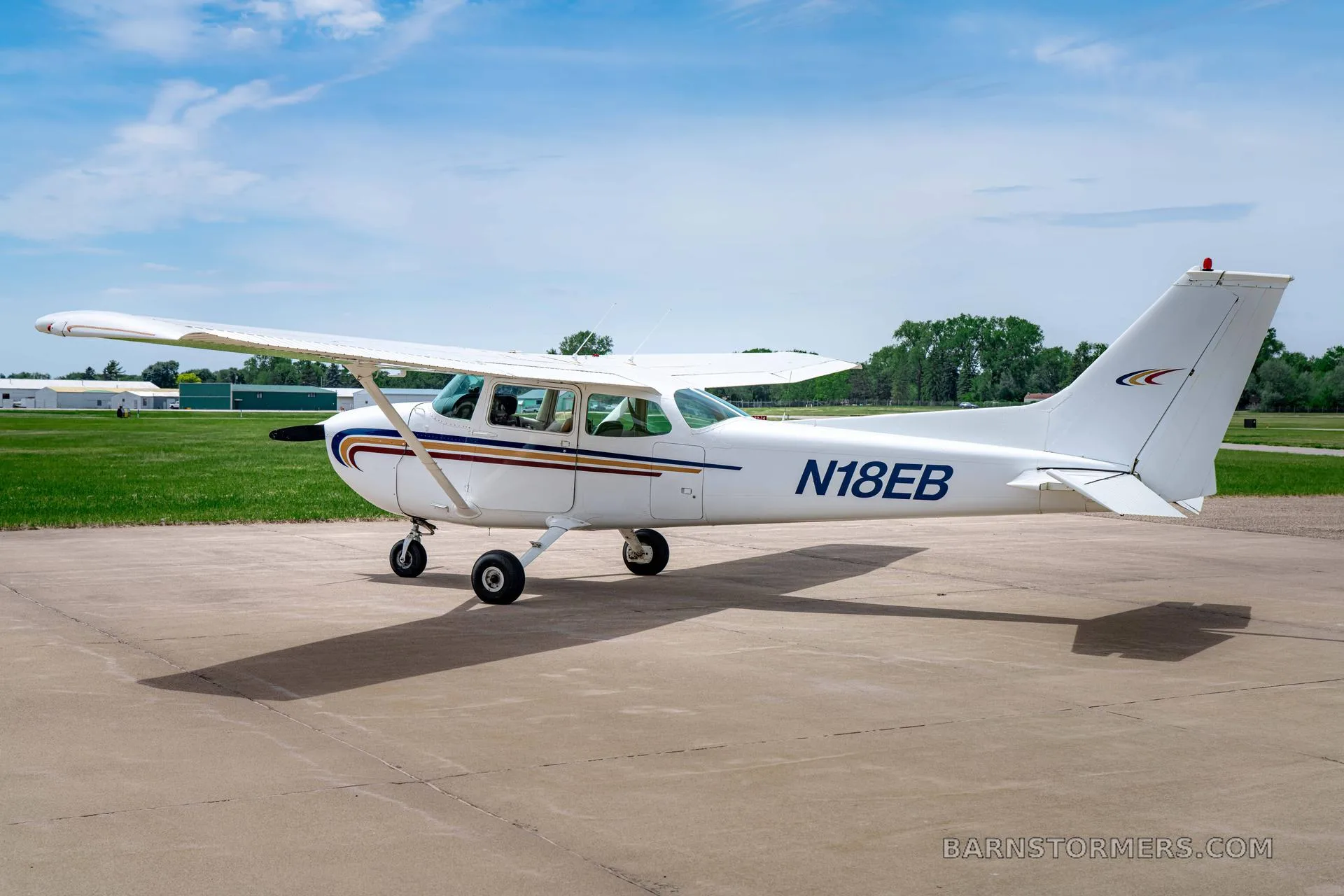 A white Cessna 172 (N18EB) with blue and gold stripes is parked on a tarmac, viewed from the rear-right.