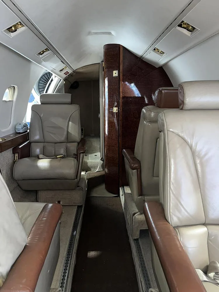A view down the aisle of a private jet cabin with beige leather seats and a wood-paneled bulkhead.