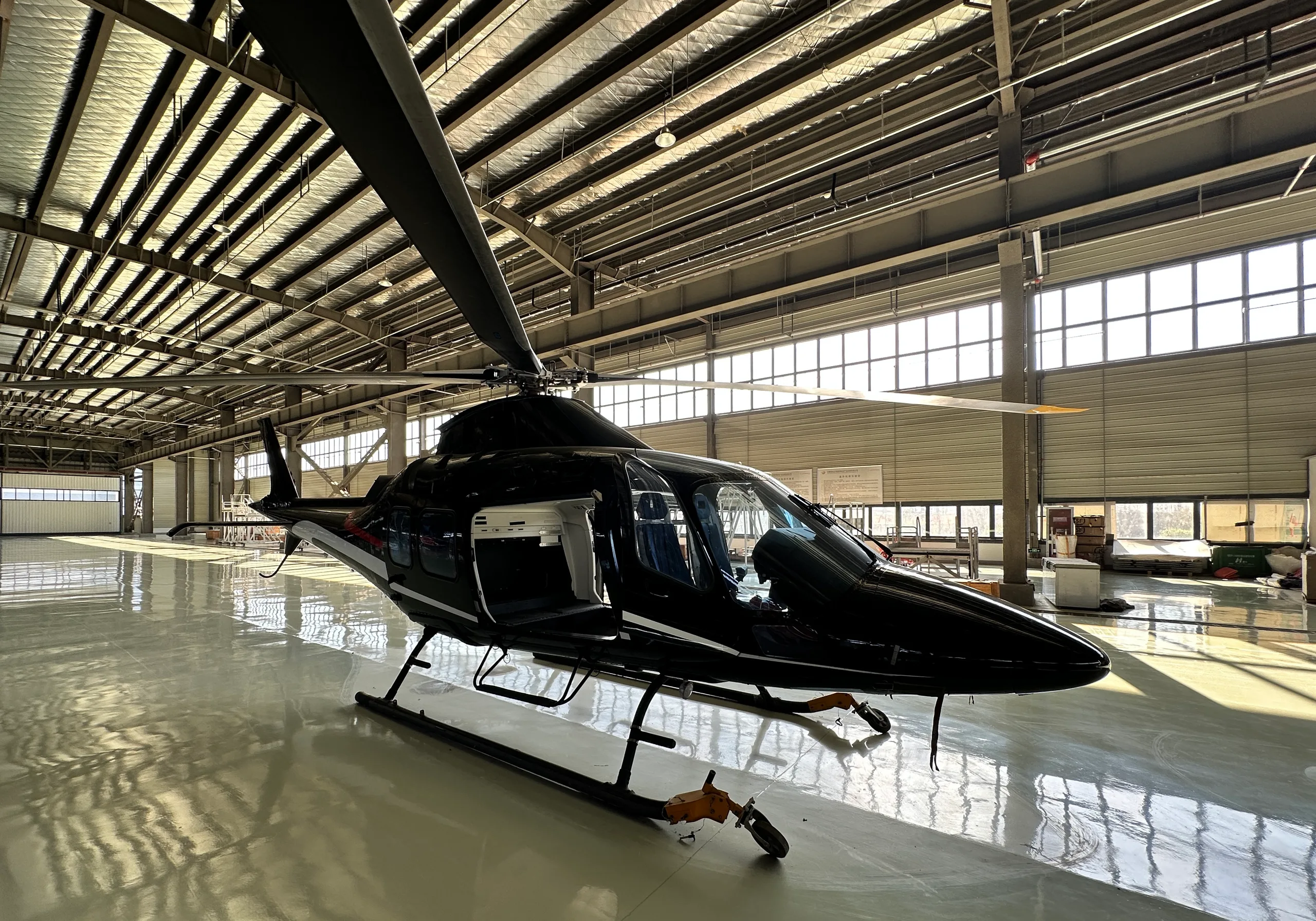 A black helicopter with white and red stripes, seen from a low front angle, parked in a brightly lit hangar with its door open.