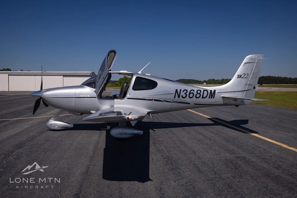 A silver Cirrus SR22 Turbo aircraft (N368DM) with its doors open, parked on an asphalt runway under a clear blue sky.