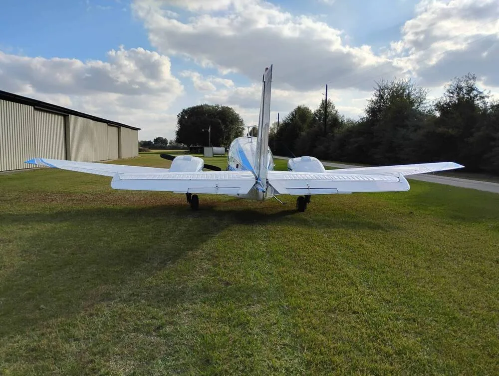 The rear view of a white Beechcraft Baron N62BB with blue stripes, parked on a grassy field near a hangar.