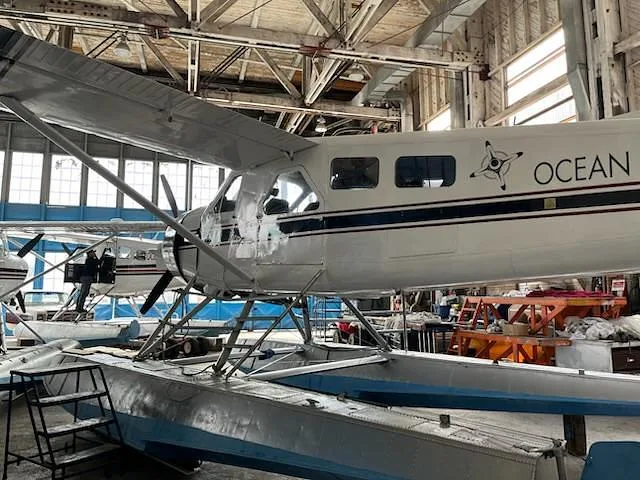 The side view of a white De Havilland Beaver seaplane with "OCEAN PACIFIC AIR" markings is shown inside a hangar, with another aircraft in the background.
