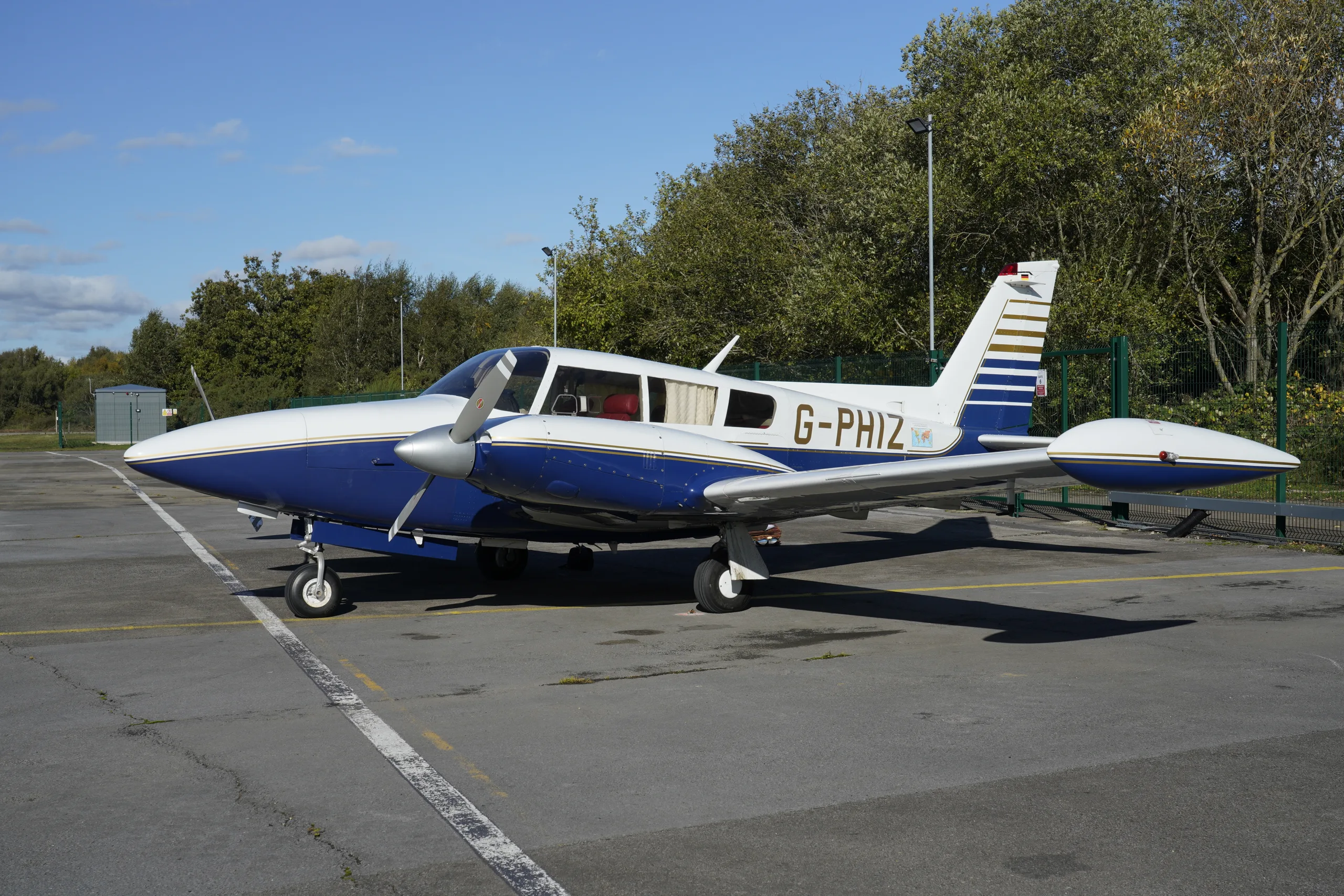 A blue and white single-engine propeller aircraft, registered G-PHIZ, is parked on an asphalt tarmac under a clear sky.