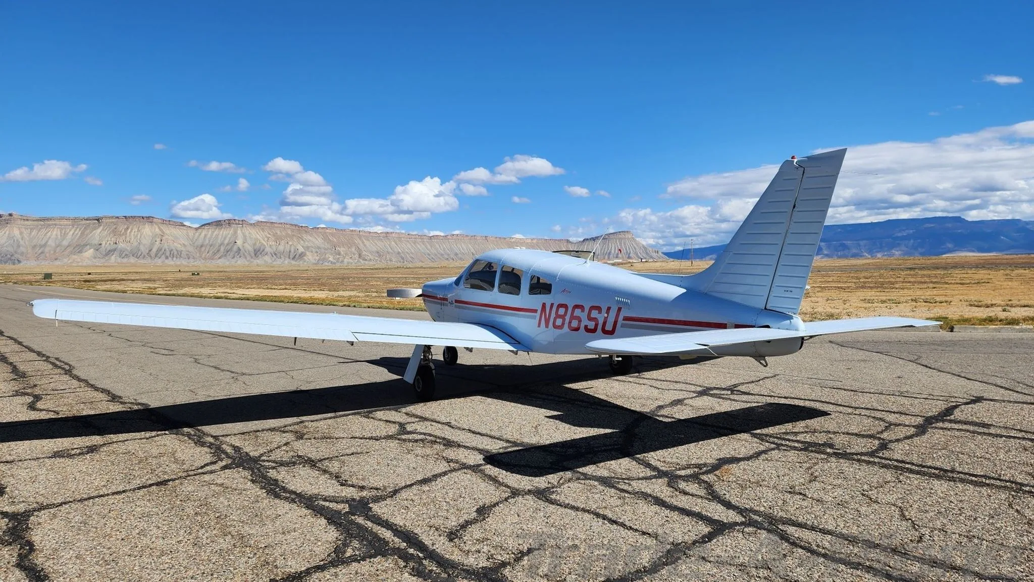 A white Piper aircraft with red stripes and tail number N86SU is parked on an asphalt runway with mountains in the background under a blue sky.