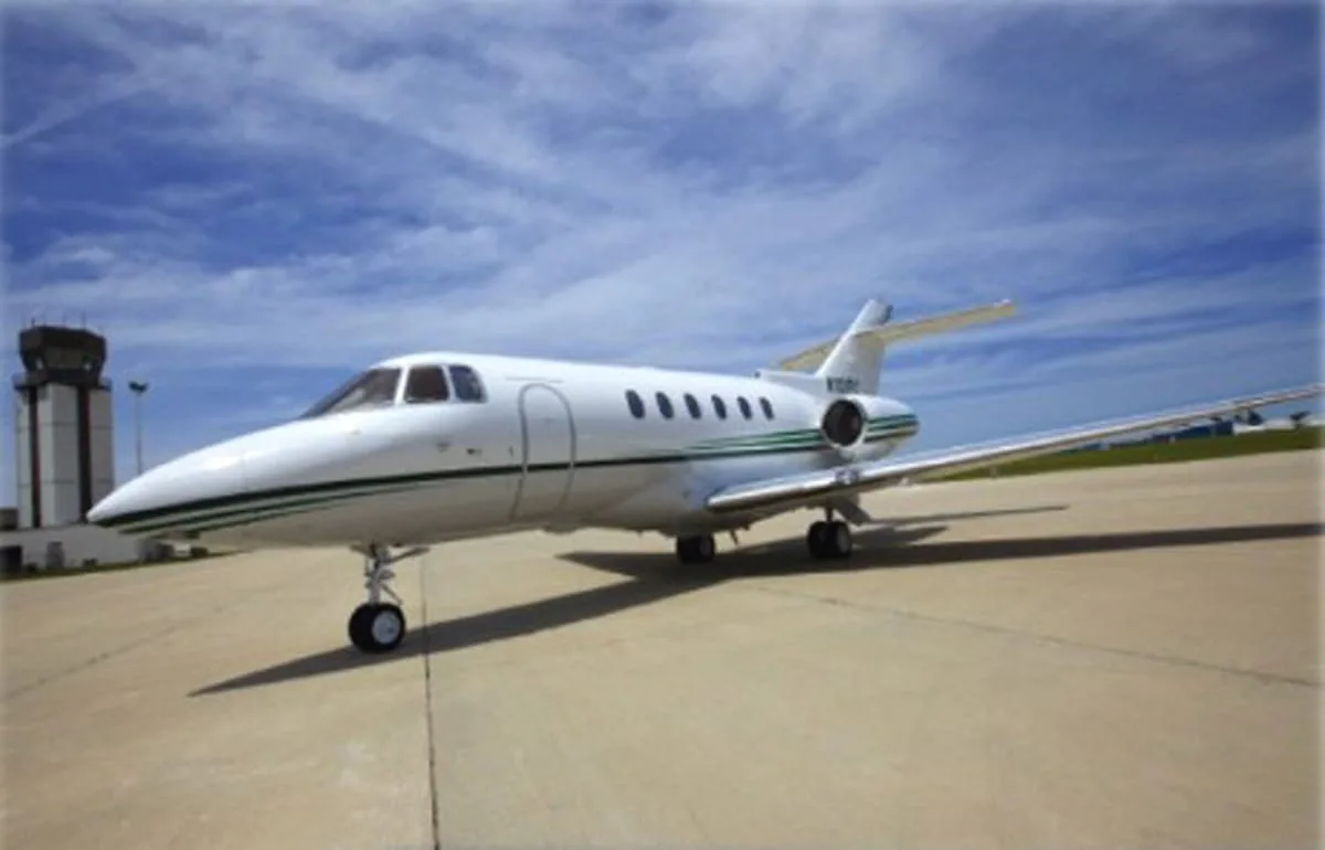 A white business jet with a green stripe on the tarmac, with an air traffic control tower in the background.