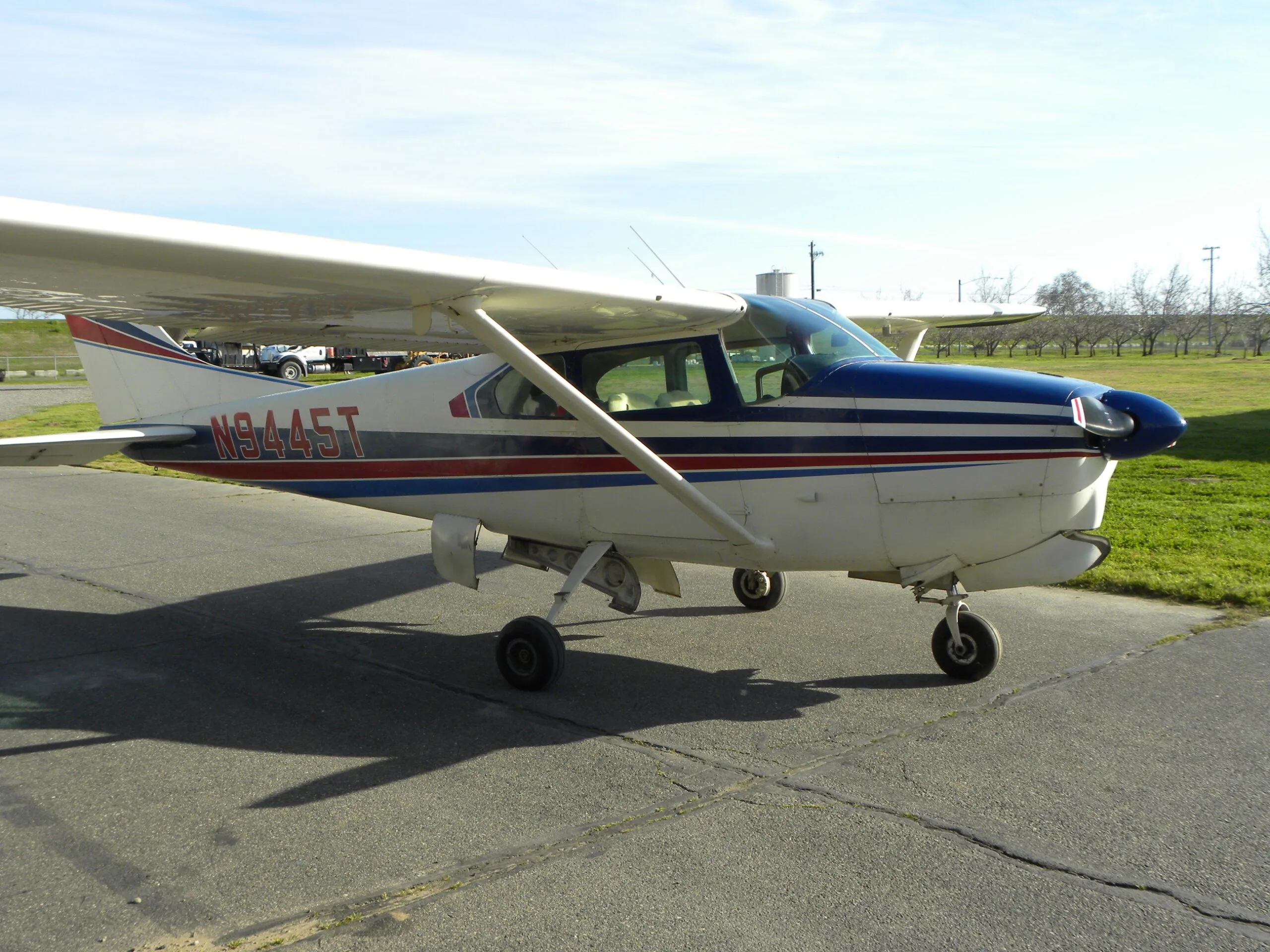 A side view of a white Cessna 182 with blue and red stripes, registration N9445T, on a tarmac.
