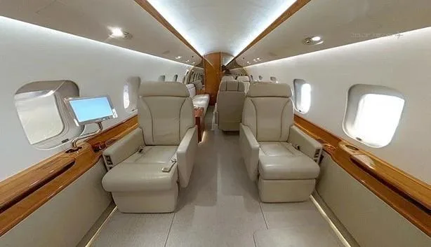 A luxurious aircraft cabin with two rows of beige leather seats facing each other, a small screen on the left, and polished wood accents.