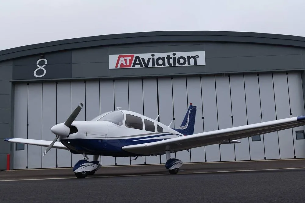 A white and blue single-engine aircraft, likely a Piper Cherokee, parked in front of an AT Aviation hangar.