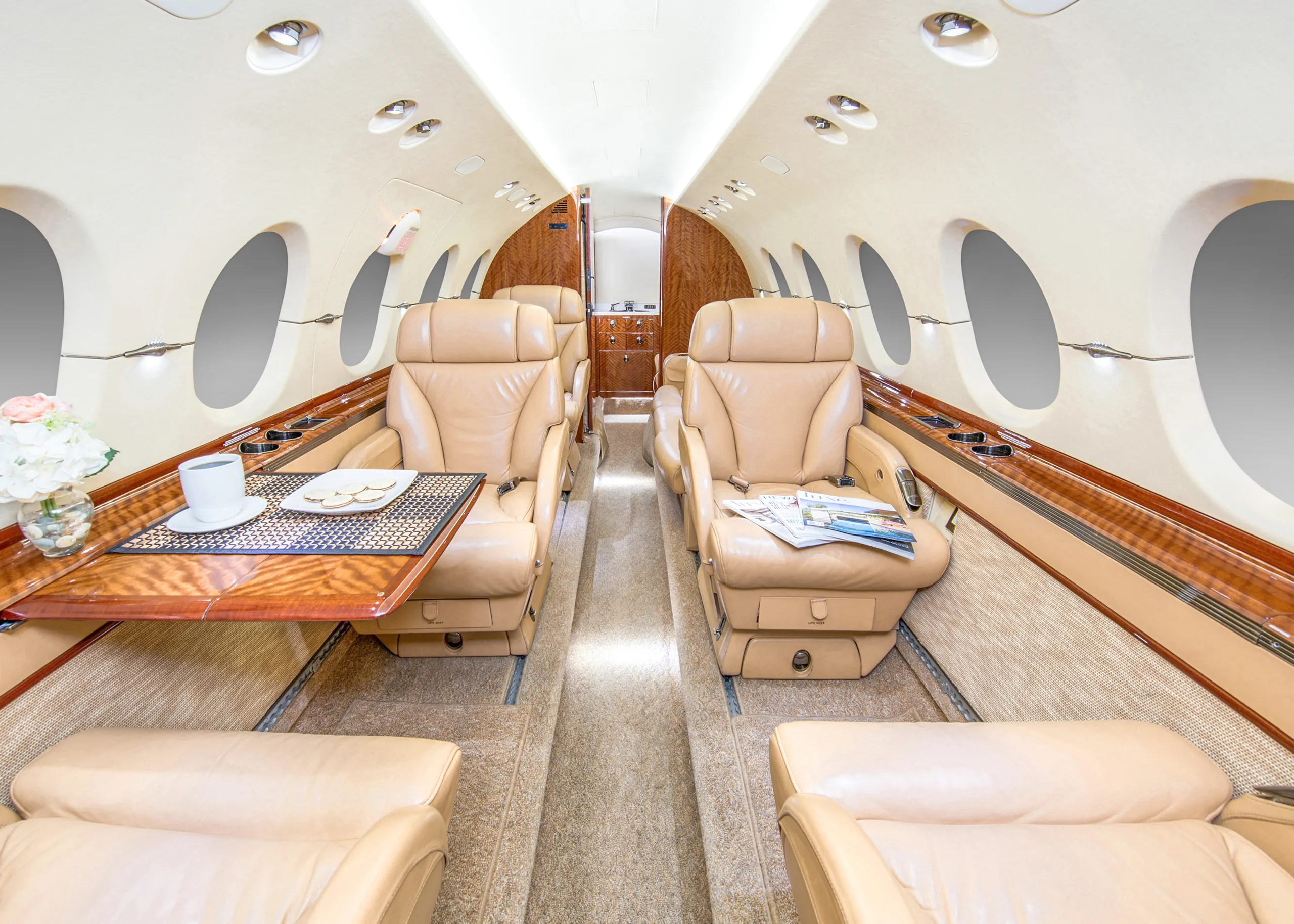 The luxurious interior cabin of a private jet with tan leather seats, a wooden table set with a cup and cookies, and large oval windows.