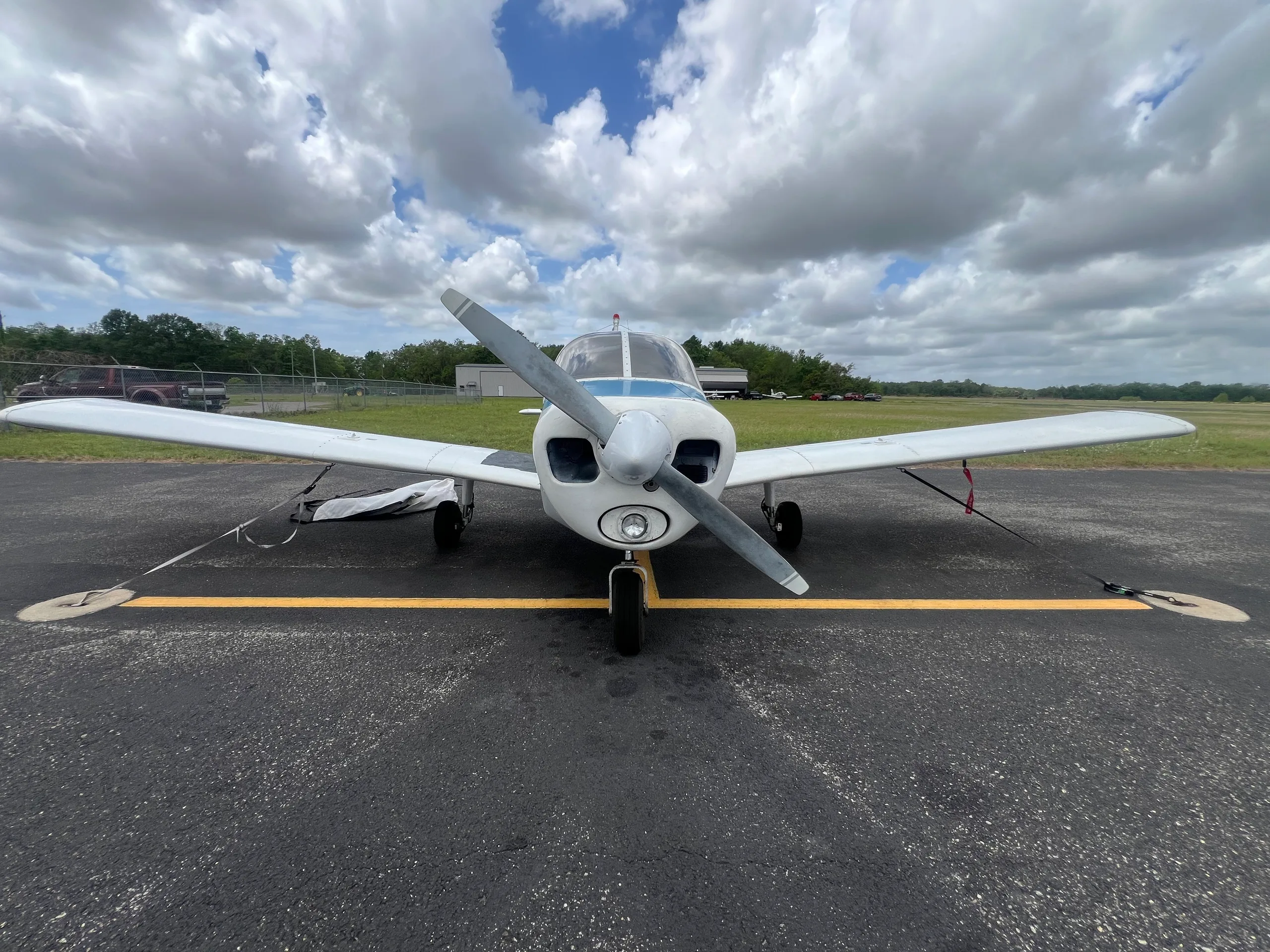 Front view of a white and blue Piper Cherokee 140 parked on an asphalt ramp under a cloudy sky.