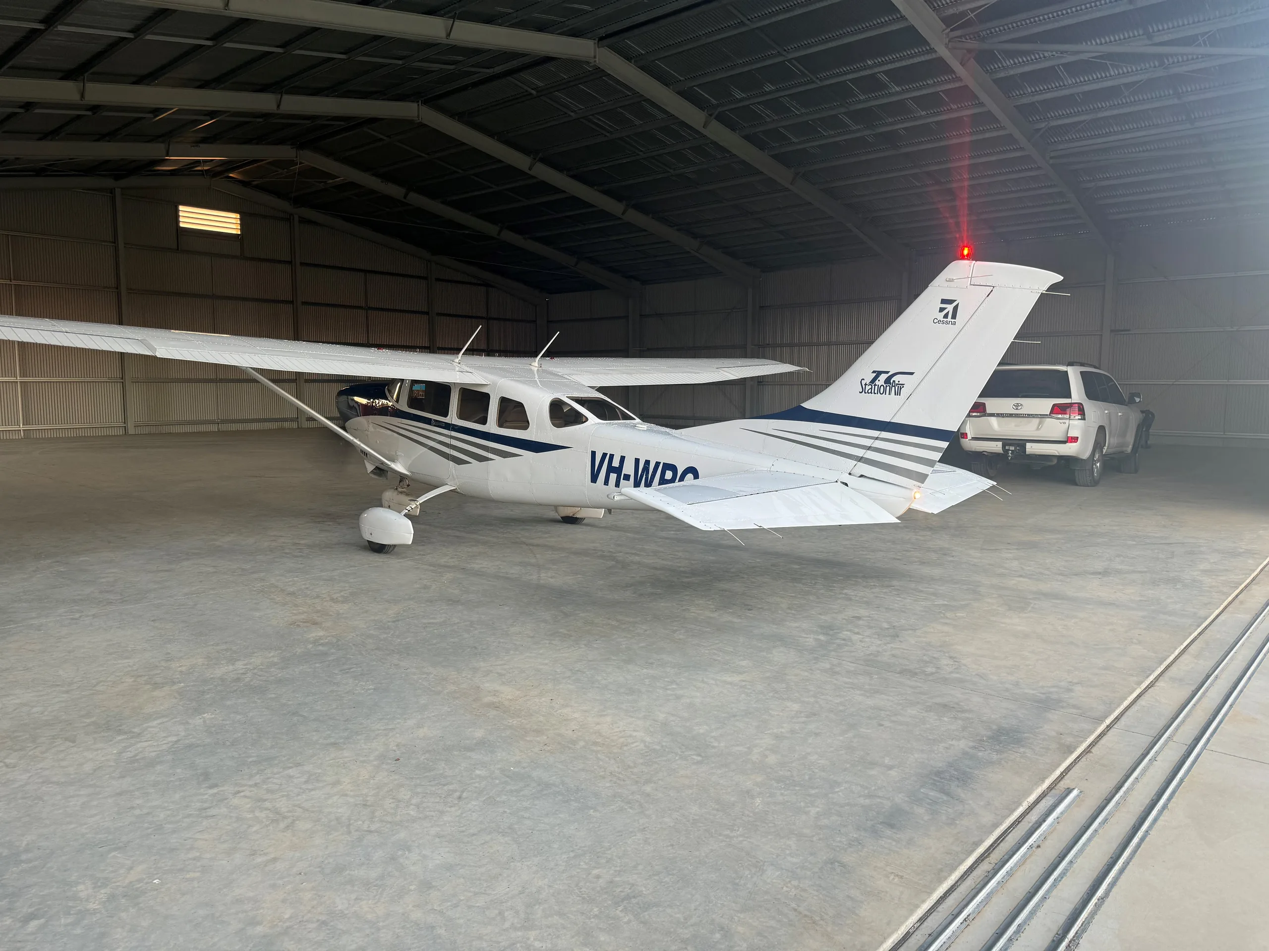 A white Cessna 210 with blue stripes, registration VH-WPC, parked inside a large metal hangar next to an SUV.