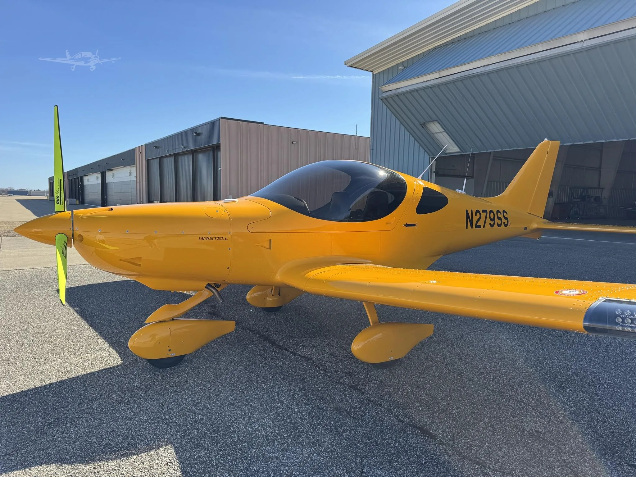 A bright yellow Bristell light sport aircraft, registration N279SS, parked on an asphalt ramp in front of hangars under a clear blue sky.