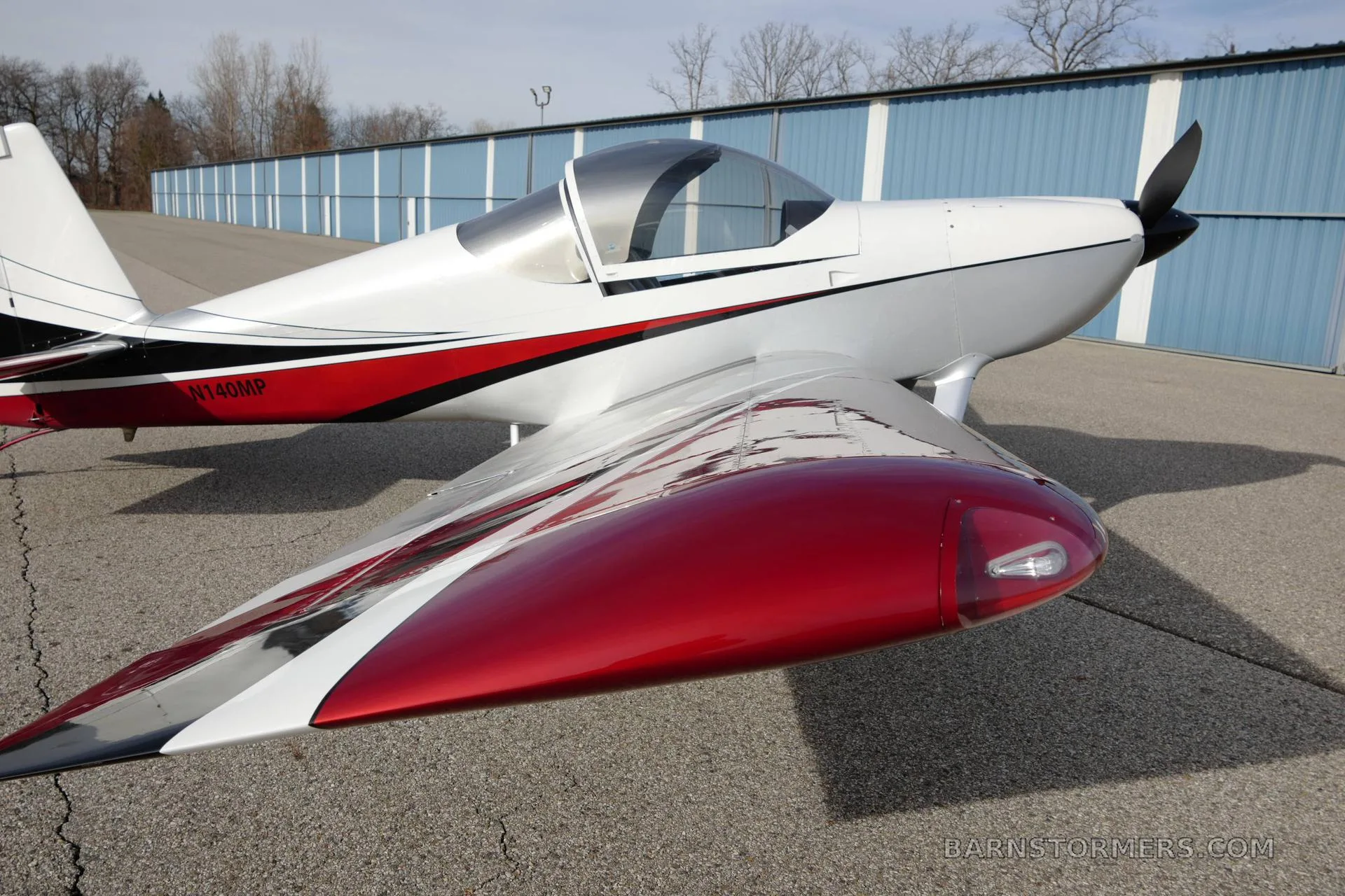 A white and red Vans RV-140MP aircraft with a shiny red wing, parked next to a blue hangar.