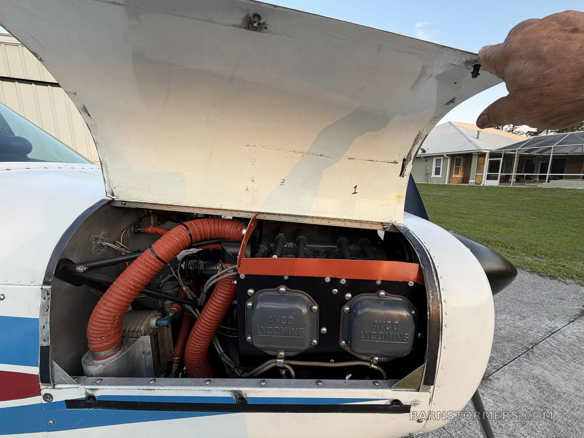A hand holds open the engine cowling of a white aircraft, revealing the Avco Lycoming engine with red hoses and components, with a house in the background.