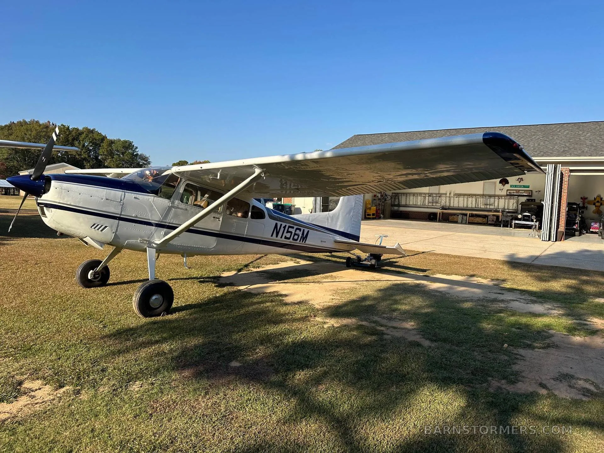 A white and blue Cessna 180 aircraft parked on a sunny grassy field in front of an open hangar.