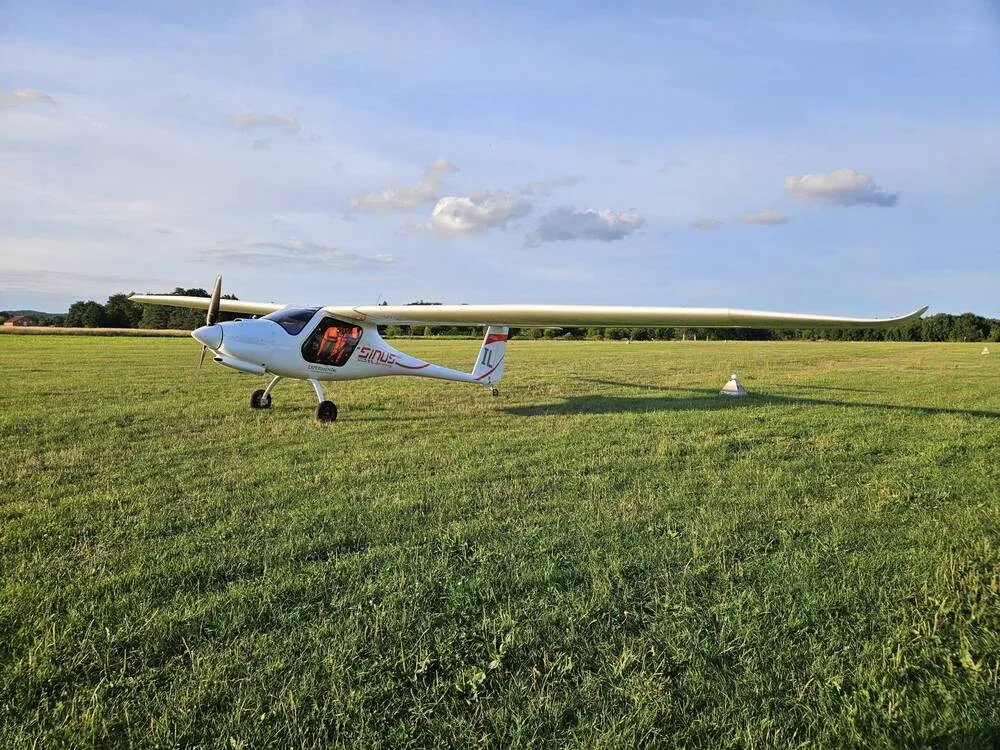 A white Pipistrel Sinus aircraft with long wings is parked on a wide grassy airfield under a blue sky.