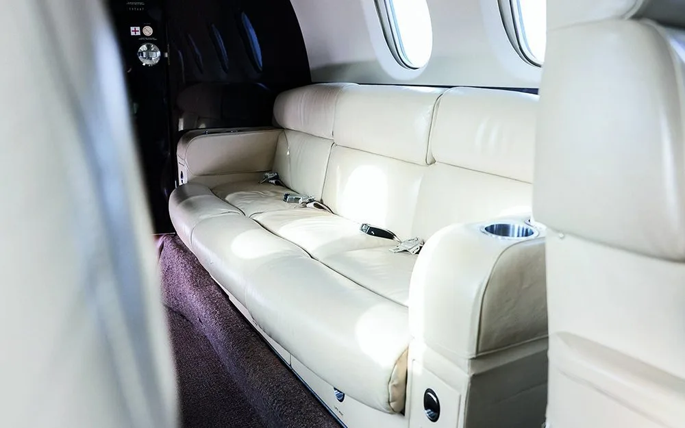 A cream-colored leather three-seat sofa with seatbelts and a cup holder in the aircraft cabin.