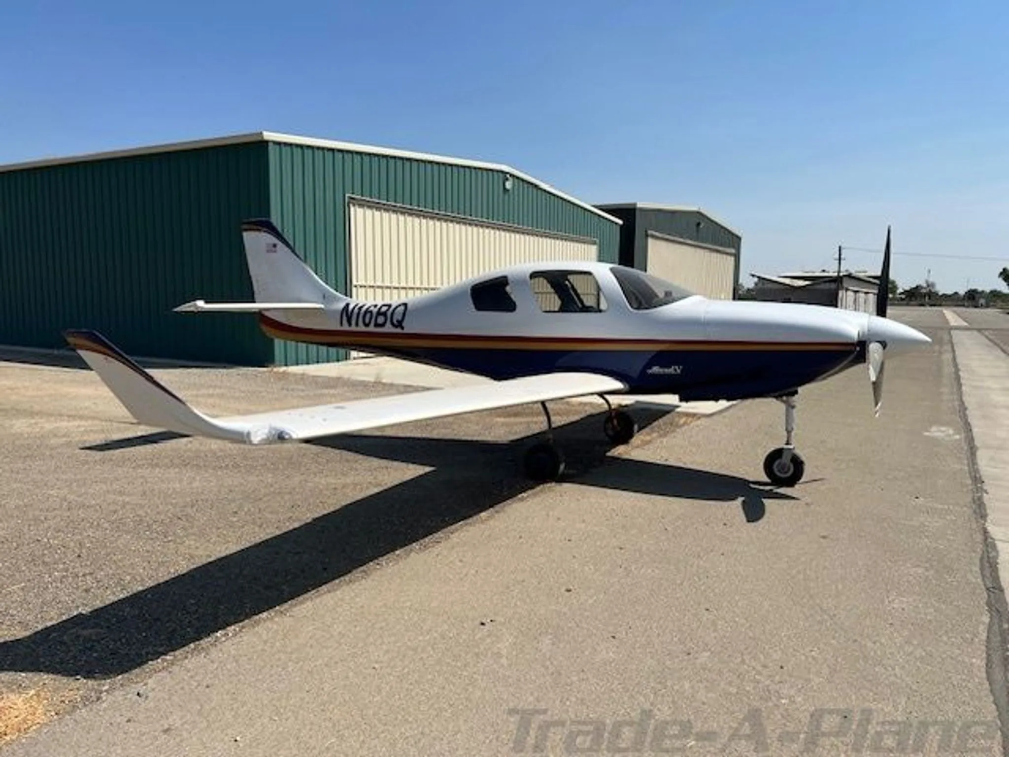 A white, blue, and gold single-engine aircraft, N16BQ, is parked on an airfield next to a green hangar under a clear sky.