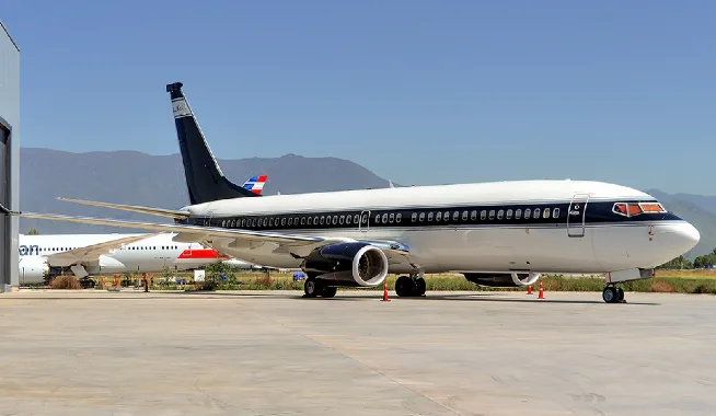 A Boeing 737 BBJ in a dark blue and white livery parked on an airfield with mountains in the background.