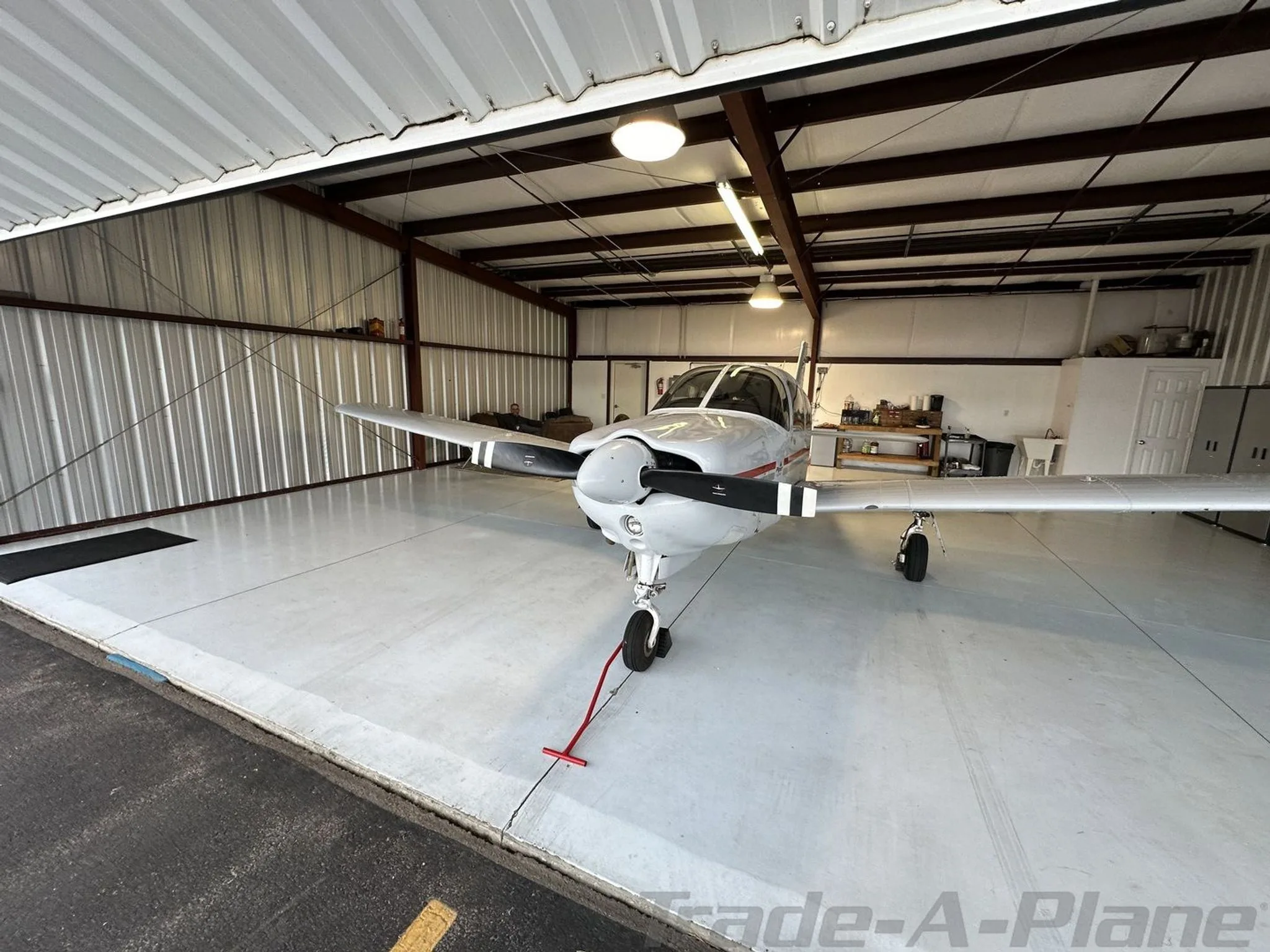 A white Piper aircraft with a black propeller is parked inside a metal hangar, viewed from a slight angle.