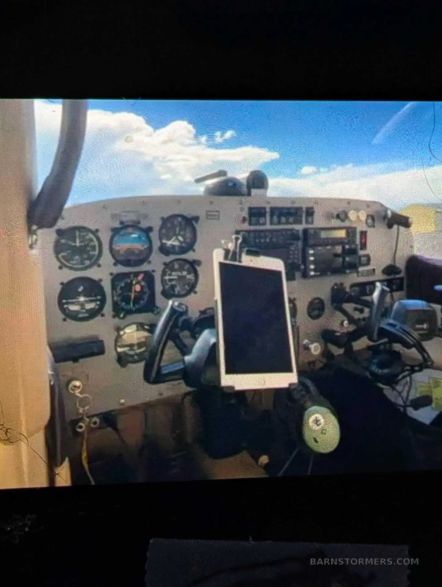 The cockpit of a small aircraft, showing the instrument panel with analog gauges and a tablet mounted on the yoke.